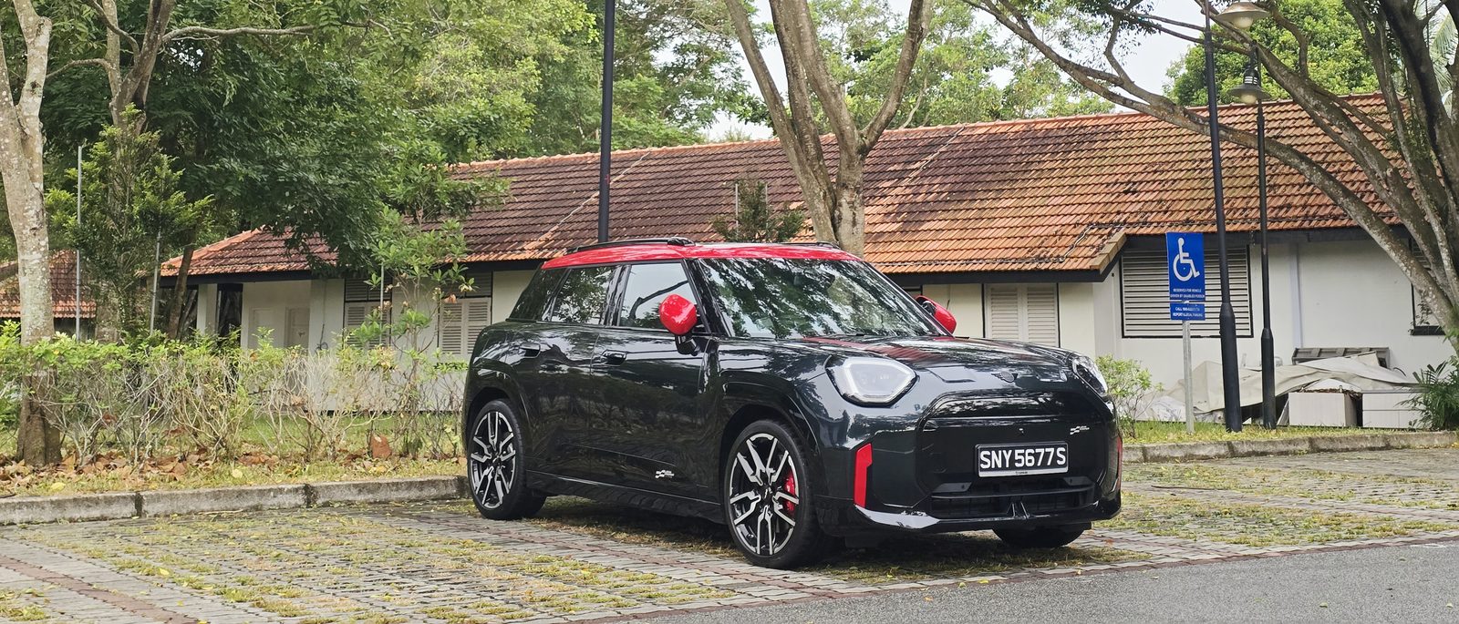 MINI JCW Aceman E in British Racing Green with a red roof, front three-quarter view parked in front of a colonial bungalow in Singapore