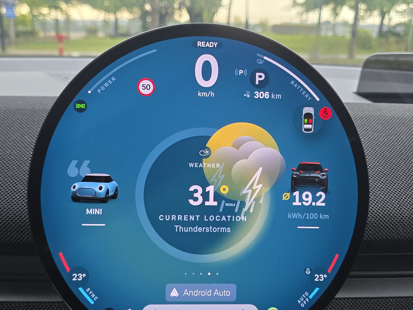 The MINI JCW Aceman E round display in teal-blue Core mode, showing the weather, the animated MINI assistant, 306 km of range and 19.2 kWh per 100 km on the trip computer