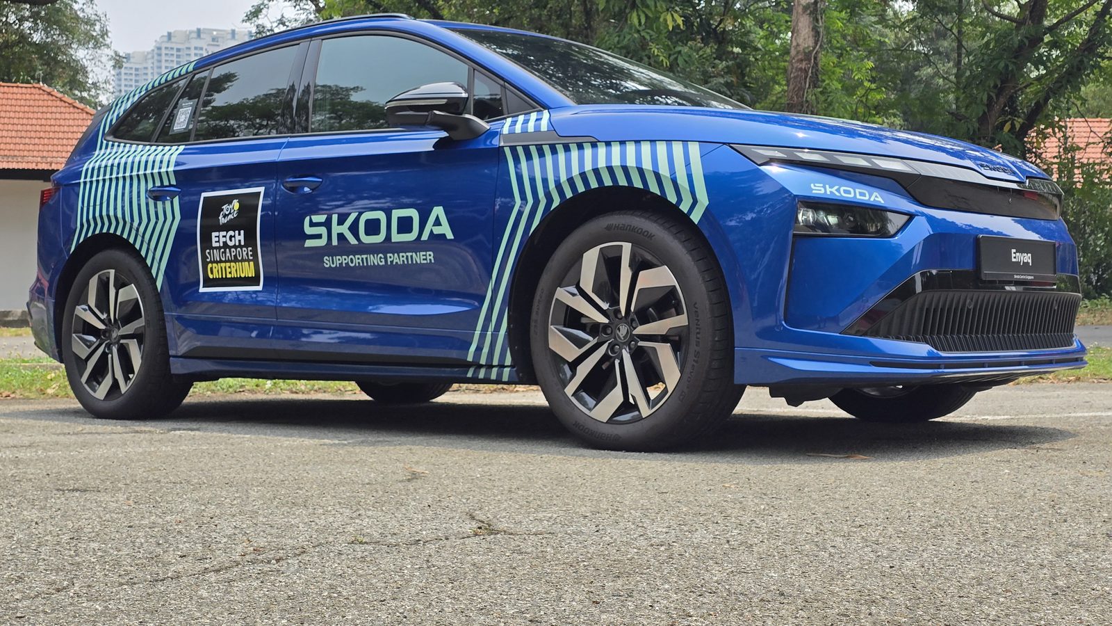 Race Blue Skoda Enyaq Sportline, front three-quarter view in a tree-lined carpark in Singapore, wearing a Tour de France EFGH Singapore Criterium wrap