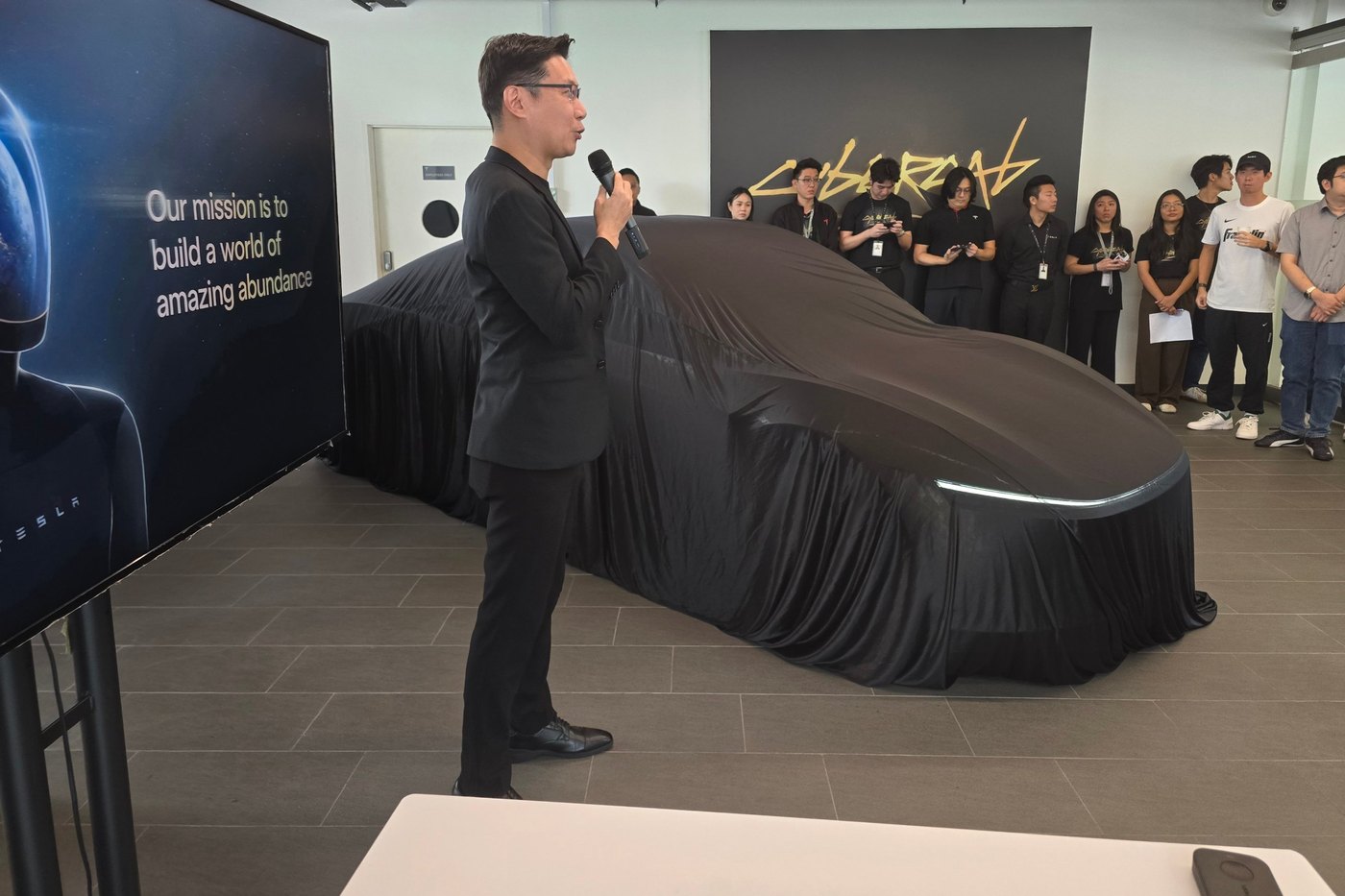 The Tesla Cybercab under a black cover at its Singapore reveal, with a Tesla Singapore spokesperson at the microphone